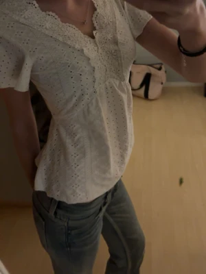 Vit blus med spetsdetaljer - Säljer en söt vit blus med broderade hålmönster och spets längs v-ringningen. Blusen har korta ärmar och en lätt utsvängd passform som ger en romantisk vibe. Perfekt till jeans eller kjol för en fräsch look. Använd 1 gång! 💗💗
