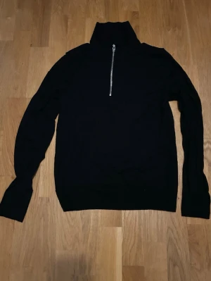 Svart stickad tröja med half zip - Svart stickad tröja med hög krage och halv dragkedja framtill. Långärmad modell som passar perfekt till kyligare dagar. Enkel och stilren design som funkar till det mesta.