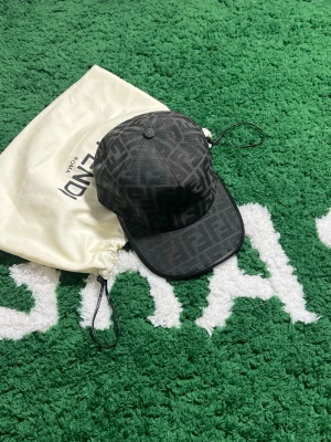 Svart Fendi keps med monogram - Säljer en svart keps från Fendi med det klassiska FF-monogrammet över hela kepsen. Kepsen har böjd skärm och justerbar rem bak. Materialet är slitstarkt och har en snygg mörk ton som passar till det mesta. Perfekt accessoar för dig som gillar exklusiva detaljer.