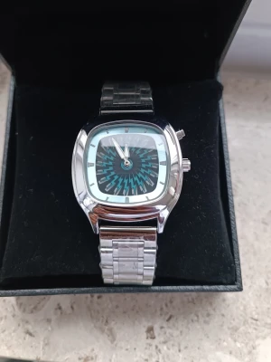 Silverklocka "Electric Zigzag" – Unik Urtavla med Blixtmönster ⚡ - Säljer en klocka som verkligen sticker ut med sin "elektriska" urtavla! Boetten är fyrkantig med rundade hörn (tv-form) och har ett häftigt mönster av blå och svarta blixtar/sicksack. Design: Retro-futuristisk "Pop Art"-stil. Påminner om 80/90-tals estetik. Detaljer: Silverfärgad länk och boett i högglans.  Skick: Helt ny och oanvänd (plasten sitter kvar på länken). Funktion: Analog tidsvisning. 