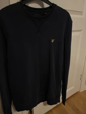 Mörkblå sweatshirt från Lyle & Scott - Mörkblå sweatshirt från Lyle & Scott med klassisk rund halsringning och gul broderad logga på bröstet. Tröjan har ribbade muddar vid ärmslut och nederkant samt är långärmad. Perfekt för en clean och stilren look.