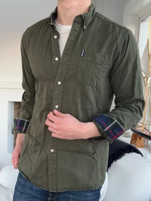 Grön skjorta från Barbour - Snygg grön skjorta från Barbour med klassisk knappslå och bröstficka. Insidan av manschetterna har rutigt tyg i powerful färger som blått, grönt och rött. Skjortan har lång ärm och är tillverkad i mjuk bomull med en tailored fit.