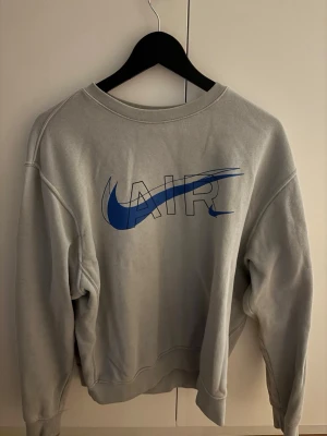 Grå Nike AIR sweatshirt  - Säljer en stilren grå sweatshirt från Nike med stor blå AIR-logga och swoosh på bröstet samt liten logga på ryggen. Tröjan har rund halsringning, långa ärmar och är gjord i mjuk bomullsblandning. Perfekt för dig som gillar sportig och clean stil.