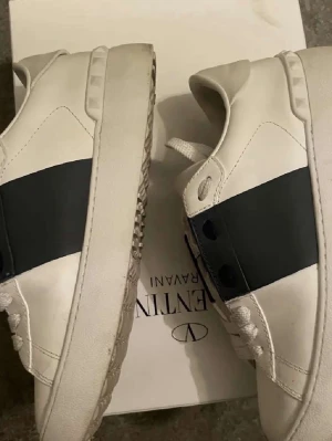 Valentino skor - Säljer ett par vita Valentino sneakers med bred svart rem över mitten och coola nitar på sulan. Skorna har rund tå, vita snören och är tillverkade i skinn. Perfekta för dig som vill ha en clean men ändå lyxig look. Pris kan diskuteras 