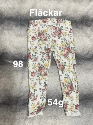 Blommiga leggings från Newbie - Supersöta vita leggings från Newbie med blommigt mönster i rosa, gul och grönt. Byxorna har elastisk midja och spetsdetalj vid bensluten. Perfekta för dig som gillar färg och mönster!🌸 Fläckar. Storlek 98