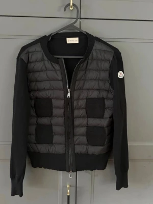 Svart Moncler Cardigan  - Säljer en svart quiltad bomberjacka från Moncler. Jackan har puffad front i dun och stickade ärmar samt rygg. Två fickor framtill och dragkedja. Klassisk Moncler-logga på ärmen. Snygg och stilren modell som passar till mycket.