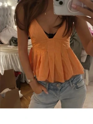 Randig orange linnetopp med v-ringning - Säljer en superfin orange linnetopp med smala vita ränder och djup v-ringning. Toppen har en figurnära överdel och ett utställd, veckad nederdel som ger en snygg siluett. Ärmlös modell med tunna axelband, perfekt för varma dagar.