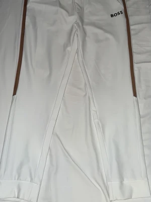 Vita BOSS mjukisbyxor - Säljer ett par vita mjukisbyxor från BOSS med bruna och svarta ränder längs sidorna. Byxorna har elastisk midja och sitter väldigt fint. Perfekta för en chill och sportig look. Materialet känns mjukt och stretchigt. Ordniariepris är 1700 kronor men nu säljs byxorna för 900