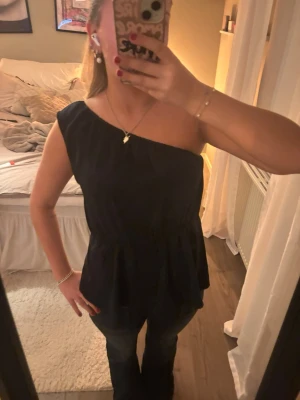 Svart oneshoulder topp med resår - Snygg mörkblå topp med oneshoulder-design och resår i midjan som ger en fin form. Toppen är ärmlös på ena sidan och har en lös passform nedtill. Perfekt att styla med jeans eller kjol för en trendig look.