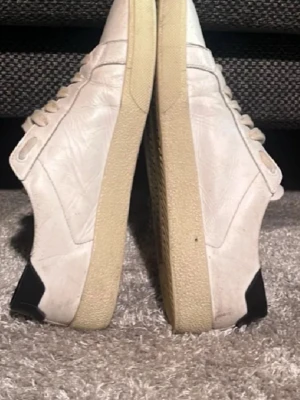 Saint Laurent vita sneakers i skinn - Säljer ett par vita Saint Laurent sneakers i skinn med handskriven logga på sidan. Skorna har beige sula, svarta detaljer vid hälen och klassisk snörning. Tillverkade i Italien och har en stilren, tidlös look som passar till många outfits. Kom med prisförslag. 