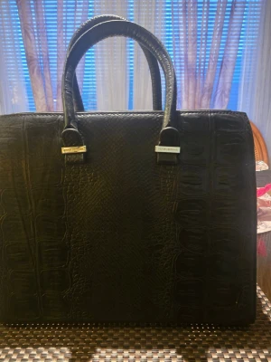 Svart handväska från Victoria Beckham - Snygg svart handväska från Victoria Beckham i strukturerat skinn med krokodilpräglat mönster. Väskan har två robusta handtag, guldfärgade metalldetaljer och ett rymligt innerfack med ficka. Perfekt för dig som vill ha en stilren och lyxig accessoar.