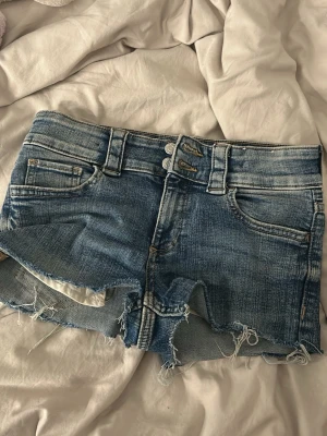 Blå jeansshorts från H&M - Säljer ett par blå jeansshorts från H&M med låg midja och råa, fransiga kanter. Shortsen har klassisk femficksdesign, dubbla knappar framtill och slitna detaljer för en avslappnad look. Perfekta för sommardagar och festivaler.