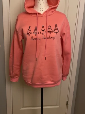 Rosa hoodie med julmotiv och huva - Säljer en rosa hoodie med huva och dragsnören. På bröstet finns ett tryck med fem stiliserade julgranar och texten 'Happy Holidays!'. Hoodien har långa ärmar och ribbade muddar. Perfekt för dig som vill ha en mysig och festlig look under vintern.