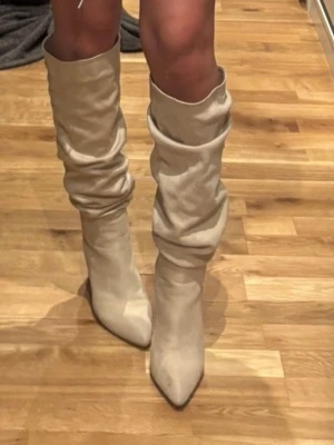 Beiga höga stövlar med spetsig tå - Snygga höga stövlar i beige mocka med slouchy passform och spetsig tå. Stövlarna har en låg klack och ger en trendig look till din outfit. Perfekta för dig som vill sticka ut med stilrena boots.
