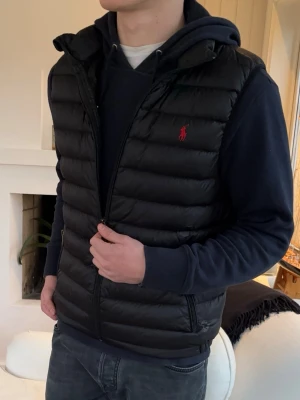 Svart dunväst från Ralph Lauren - Svart dunväst från Ralph Lauren med röd broderad logga på bröstet. Västen har dragkedja framtill, två sidofickor med dragkedja och en sportig, quiltad design. Perfekt att slänga över en hoodie för en chill look. Finns en lagning på västen
