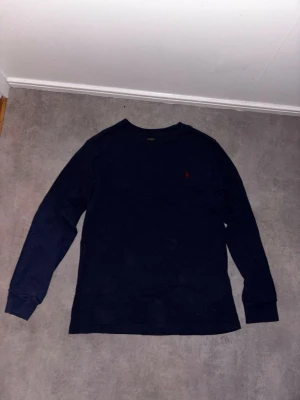 Mörkblå långärmad tröja Polo Ralph Lauren - Mörkblå långärmad tröja från Polo Ralph Lauren med klassisk röd logga broderad på bröstet. Tröjan har rund halsringning och är tillverkad i mjukt bomullsmaterial. Perfekt för en stilren och avslappnad look.Stilren vit T-shirt från Lacoste med det ikoniska krokodilmotivet och texten Lacoste Paris framtill i mörkblått. Klassisk och tidlös design som passar till det mesta. Tillverkad i mjuk och bekväm bomull, perfekt både till vardag och mer uppklädda outfits och tröjorna är köpta från zalando.