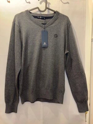 Grå stickad tröja från Morris - Klassisk grå stickad tröja från Morris med V-ringning och diskret broderad logga på bröstet. Tröjan har långa ärmar och ribbade muddar vid ärmslut och nederkant. Perfekt för lager-på-lager och enkel att matcha.