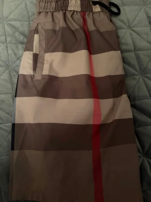 Burbery shorts  - Snygga badbyxor med klassiskt rutigt mönster i beige, brunt, svart och rött. De har elastisk midja med svart dragsko och sidofickor. Perfekta för stranden eller poolen och ger en stilren look. Storlek L men liten i storlek passar m
