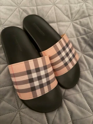 Rutiga tofflor från Burberry - Snygga tofflor från Burberry med klassiskt rutig mönster i beige, svart, vitt och rött. Tofflorna har en bred rem över foten och svart sula. Perfekta för en chill och trendig look.