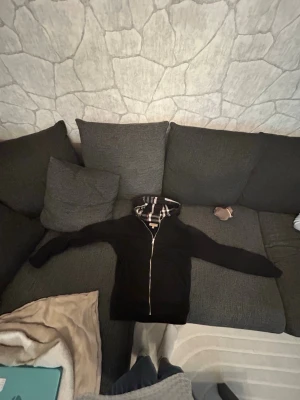 Svart hoodie från Burberry med dragkedja - Säljer en svart hoodie från Burberry med dragkedja framtill och klassiskt rutigt foder i huvan. Hoodien har långa ärmar och en stilren look som passar perfekt till streetwear. Materialet känns mjukt och bekvämt, och detaljerna är riktigt snygga.