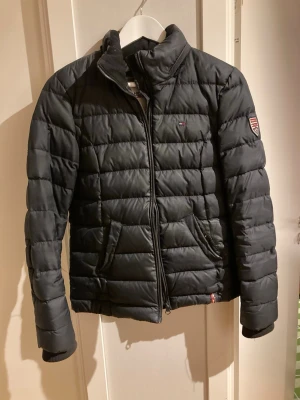 Svart pufferjacka från Hilfiger Denim - Snygg svart pufferjacka från Hilfiger Denim med quiltad design och hög krage. Jackan har dragkedja framtill, två sidofickor och ett broderat märke på ärmen. Perfekt för kalla dagar och riktigt stilren look.