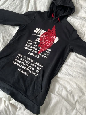 Svart Nike Air Jordan hoodie - Snygg svart hoodie från Nike Air Jordan med stor vit och röd print framtill. Hoodien har huva med röda snören och känguruficka. Materialet är mjukt och skönt, perfekt för en chill look. Stora loggor och text på både engelska och franska ger en unik vibe.