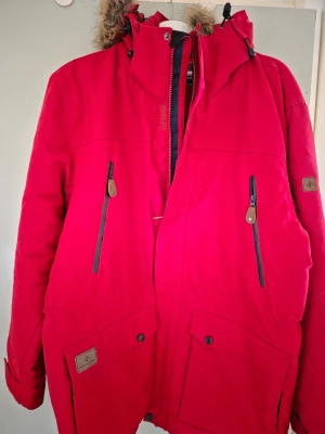 Röd vinterjacka med fuskpäls - Snygg röd parkasjacka med huva och fuskpälsdetalj. Jackan har dragkedja framtill, flera fickor med både dragkedja och knappar samt justerbara muddar. Perfekt för kalla dagar och har en klassisk parkaslook.