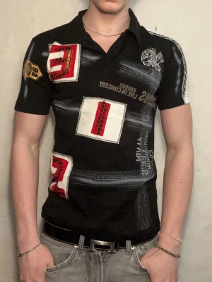 Svart mönstrad pikétröja  - Cool svart pikétröja från Ed Hardy med tryck och broderade detaljer i rött, vitt och gult. Tröjan har korta ärmar, krage och flera grafiska mönster och siffror på både fram- och baksida. Perfekt för dig som gillar streetwear med attityd.