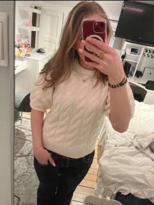 Vit kabelstickad tröja med puffärm - Säljer en vit kabelstickad tröja med korta puffärmar och rund hals. Tröjan har ett klassiskt stickat mönster och ribbade muddar vid ärmslut och nederkant. Perfekt för lager-på-lager och snygg till både jeans och kjol.