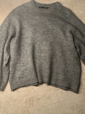Grå stickad tröja  - En enkel och stilren grå stickad tröja med rund hals. Tröjan har ribbstickade muddar vid ärmslut och nederkant. Perfekt för lager-på-lager och passar till det mesta.