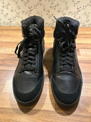 Svarta höga sneakers från Puma - Snygga svarta sneakers från Puma med högt skaft och snörning. Skorna har en mjuk, fluffig insida och detaljer i både skinn och mocka. Puma-logga syns på hälen och plösen. Perfekta för dig som gillar sportig och stilren look.