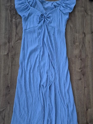 Ljusblå långklänning med volang från lager 157 - Säljer en ljusblå långklänning med volangdetaljer vid axlarna och knyt framtill. Klänningen har en mjuk, lätt textur och är perfekt för varma dagar. Den har en feminin siluett och är tillverkad i ett luftigt material.
