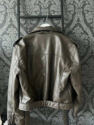 Brun skinnjacka från Gina Tricot - Snygg brun skinnjacka från Gina Tricot med bred krage, axelklaffar och bälte i midjan. Jack soppar en klassisk bikerstil och är perfekt för dig som vill ha en edgy look. Tillverkad i skinnimitation och har en cool, oversized passform.