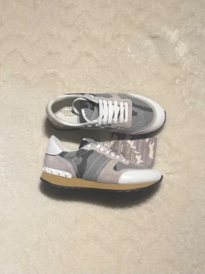Valentino Rockrunner  - Snygga Valentino Rockrunner sneakers i grått och vitt med camo-mönster. Skorna har detaljer i mocka och skinn, vita snören och en beige sula med nitar på hälen. Perfekta för dig som vill ha en unik och trendig look.