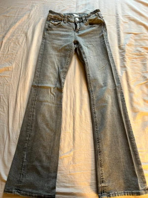 Grå bootcut jeans med fickdetaljer - Snygga grå bootcut jeans med ljus tvätt och dekorativa fickor bak. Klassisk femficksmodell med knapp och dragkedja framtill. Jeansen har en utsvängd passform och är perfekta för en avslappnad men trendig look.