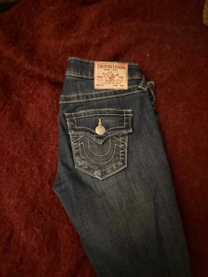 True Religion blå bootcut jeans - Säljer ett par klassiska blå jeans från True Religion med kontrastsömmar och ikonisk fickdesign med lock och knapp. Jeansen har bootcut passform och är tillverkade i slitstarkt denim. Perfekta för dig som gillar en avslappnad men snygg stil. Pris kan diskuteras 😊