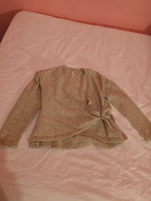 Beige omlottkofta med knyt - Mysig beige omlottkofta med knyt i sidan och långa ärmar. Koftan har en mjuk och lite melerad yta som ger en avslappnad look. Perfekt att slänga på sig över en topp för en chill stil.