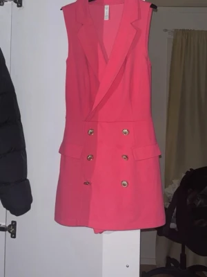 Kort rosa ärmlös klänning med knappar - Säljer en kort, ärmlös rosa shortsklänning med dubbelknäppning framtill och breda slag. Klänningen har dekorativa fickor och en figurnära passform. Perfekt för dig som vill sticka ut med en färgstark och trendig look.