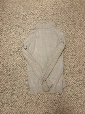 Ljusgrå stickad tröja Bondelid - Ljusgrå stickad tröja från Bondelid med halv dragkedja och hög krage. Tröjan har ribbade muddar vid ärmslut och nederkant samt en stilren, enkel design som passar perfekt till lager-på-lager. Mjuk och skön kvalitet.