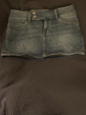 Blå jeanskjol från H&M - Säljer en blå lågmidjad jeanskjol från HM med två fickor och dubbla silverknappar. Kjolen har klassisk denimlook med slitningar och kontrastsömmar. Perfekt för dig som gillar en avslappnad och trend sopad stil.