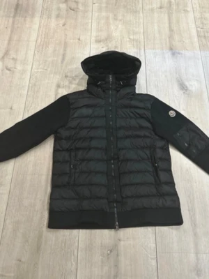 Svart cardigan  från Moncler med huva - Snygg svart cardigan  från Moncler med huva och ribbade ärmar. Jackan har dragkedja framtill, två sidofickor och en extra ficka med dragkedja på ärmen. Klassisk Moncler-logga på armen. Perfekt för kalla dagar och riktigt stilren look.