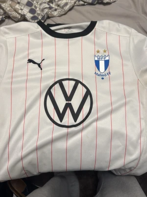 Malmö FF bortatröja Puma vit - Säljer en vit Malmö FF bortatröja från Puma med tunna röda ränder, svart krage och klubbmärke på bröstet. Tröjan har Volkswagen-logga framtill och Tillmobil-tryck på ryggen. Tillverkad i lätt och ventilerande material, perfekt för match eller träning.