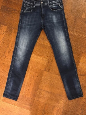 Replay Anbass jeans blå, storlek 30/32 - Snygga Replay Anbass jeans i mörkblå tvätt med slitningar och kontrastsömmar. Modellen har fem fickor, dragkedja och knapp framtill. Jeansen är raka och har en modern passform som sitter riktigt bra. Perfekt för dig som gillar stilrena och trendiga jeans. Jeansen är i storlek 30 i midjan och 32 i längden