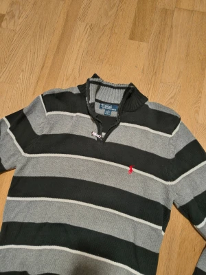 Randig stickad tröja från Polo Ralph Lauren - Snygg stickad tröja med breda svarta och grå ränder från Polo Ralph Lauren. Tröjan har halv dragkedja vid halsen, ribbade muddar och den klassiska röda logotypen broderad på bröstet. Perfekt för dig som gillar stilrena och tidlösa plagg.