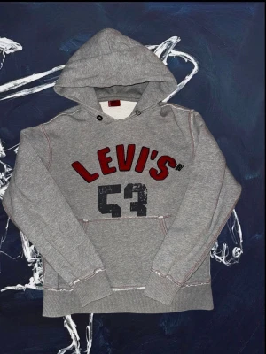 Vintage hoodie från Levi's med tryck - Snygg grå hoodie från Levi's med röd broderad logga och siffran 53 i svart tryck på bröstet. Hoodien har huva med dragsko, magficka och ribbade muddar vid ärmar och nederkant. Perfekt för en avslappnad och sportig stil.