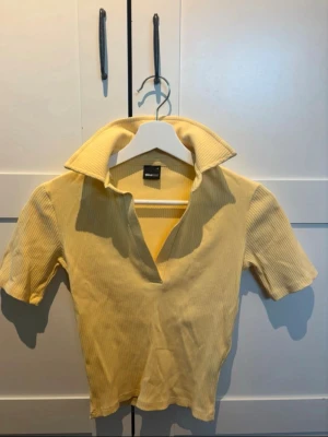 Söt gul topp från Gina Tricot! - Trendig gul ribbad topp från Gina Tricot med korta ärmar och djup v-ringning. Snygg krage och perfekt att styla med jeans eller kjol för en fräsch look.