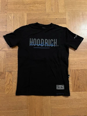 Svart Hoodrich t-shirt med tryck - Snygg svart t-shirt från Hoodrich med stort tryck i grått och blått på bröstet. Klassisk passform med korta ärmar och rund halsringning. Tillverkad i mjuk bomull och har detaljerade logotyper både fram och bak samt på ärmen.