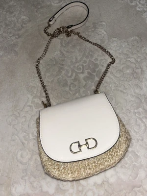 Vit och beige axelväska med kedja - Snygg axelväska  i vit och beige med guldfärgad kedjerem. Väskan har ett elegant spänne framtill och är tillverkad i skinn och flätad syntet. Perfekt för dig som vill ha en stilren och trendig accessoar.