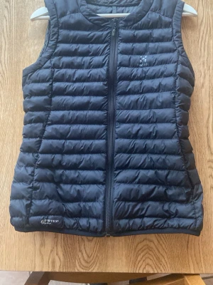 Svart dunväst Haglöfs Essens Mimic S - Säljer en svart dunväst från Haglöfs, modell Essens Mimic Vest Women. Västen har quiltad design, dragkedja framtill och är tillverkad i lätt syntetmaterial med Pertex-yttertyg. Perfekt för lager-på-lager under kyliga dagar.