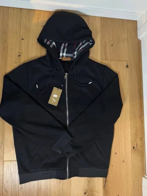 Svart hoodie från Burberry med dragkedja - Snygg svart hoodie från Burberry med dragkedja framtill och klassiskt rutigt foder i huvan. Broderad Burberry-logga på bröstet och justerbara snören i huvan. Tillverkad i mjukt bomullsmaterial för en bekväm känsla. Perfekt för en stilren och avslappnad look.